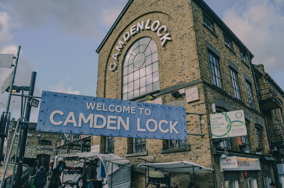 An outdoor scene at Camden Lock, featuring a large illuminated sign with bold yellow and red lettering, spanning across a metal bridge structure. Below the sign, busy street traffic includes a red dou