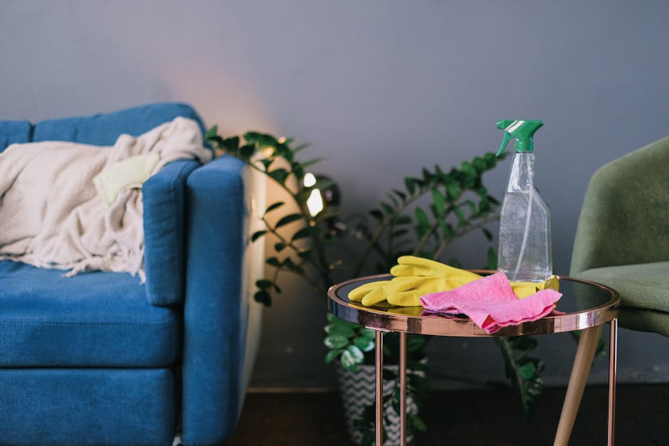 A close-up of a small, round side table in a living room, with a reflective black surface and metallic rose gold legs. On the table, there are bright yellow rubber cleaning gloves, a pink microfiber c