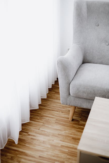 A close-up view of a living room corner featuring a light grey upholstered armchair with wooden tapered legs, positioned next to a white curtain and a window, allowing natural light to illuminate the